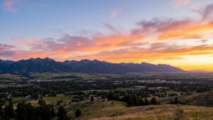 A wide landscape photograph of a winding dirt trail on a grassy hill overlooking a mountain range at sunset.