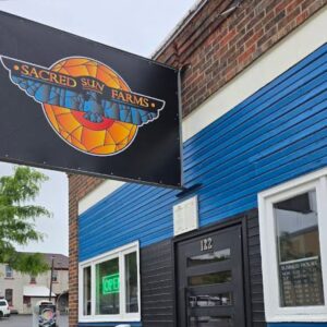 An exterior view of a storefront featuring a large black hanging sign for Sacred Sun Farms. 