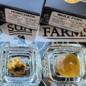 A top-down view of two small glass jars containing different cannabis concentrates, positioned next to their respective product packaging.